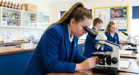 Female student in blue lab coat using microscope in science classroom laboratory for educational research and learning.