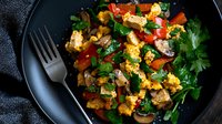 Colorful scrambled eggs with fresh vegetables and herbs served on black plate. Homemade breakfast with mushrooms, bell peppers, and parsley on dark background.