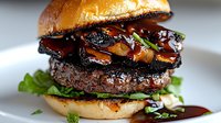 Juicy beef burger topped with glazed mushrooms and fresh greens on toasted brioche bun, drizzled with dark savory sauce. Perfect for restaurant menu photography.