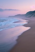Serene coastal landscape at sunset with gentle waves washing onto sandy beach beneath towering cliffs, bathed in soft pink and purple twilight hues.