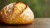 Fresh crusty artisan sourdough bread loaf with golden brown crust and flour dusting on rustic wooden surface against blurred natural background.