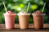 Three delicious milkshakes with whipped cream and chocolate toppings in plastic cups with straws on wooden table against blurred green background.