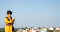 Young South Asian girl wearing traditional yellow kurta with purple dupatta, holding mobile phone outdoors against rural village landscape backdrop for cultural lifestyle and technology adoption themes.