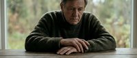 Elderly Caucasian man in green sweater sitting at wooden table looking contemplative and melancholic by window with natural light for mental health awareness campaigns.