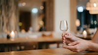 Empty wine glass held by woman's hand in upscale restaurant or bar setting with blurred elegant interior background for hospitality and dining concepts.