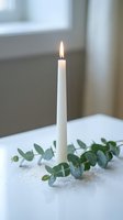 Single white taper candle burning with eucalyptus branches on white table near window creating peaceful minimalist home decor atmosphere for relaxation.