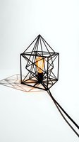 Geometric black wire cage table lamp with glowing Edison bulb casting dramatic shadows on white background, modern industrial interior decor.