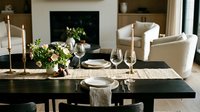 Elegant dining table set with fresh floral centerpiece, place settings, and wine glasses in modern living room with fireplace and neutral furnishings.