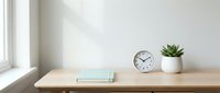 Wooden desk with clock, succulent plant, and notebook near bright window, minimalist workspace setup for home office or study area.