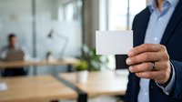 Professional man in business attire holding blank white card in modern office environment with colleagues working at desk in background, suitable for corporate branding and business communications.
