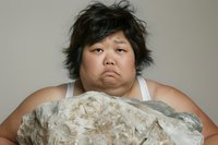 Young Asian woman with messy black hair wearing white tank top shows disappointed expression while lying on textured gray pillow against neutral background.