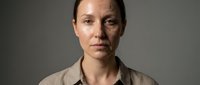 Middle-aged Caucasian woman with serious expression and bruised face wearing beige shirt against gray background for domestic violence awareness campaigns.
