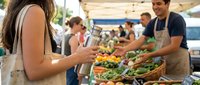 Young woman with reusable bag and bottle shopping fresh vegetables at outdoor farmers market, smiling vendor serving customers at colorful produce stand.