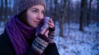Young Caucasian woman in purple knit hat and scarf drinking hot beverage from mug in snowy winter forest during twilight hour for seasonal lifestyle content.
