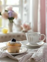 Fresh vanilla cupcake with blueberry on top and white teacup on elegant table setting with embroidered cloth napkin and soft morning light.