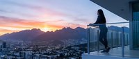 Woman sitting on modern balcony railing overlooking sprawling cityscape at sunset with mountain range in background. Urban lifestyle and aspirational living concept.