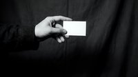 Male hand holding blank white business card, black and white photo, dark fabric background, copy space for branding mockup.