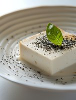 Fresh tofu block garnished with black sesame seeds and green basil leaf on ceramic plate, close-up view showing texture and minimalist presentation.