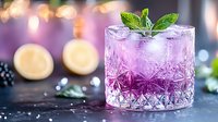 Refreshing purple cocktail in a crystal glass with ice, mint garnish, blackberries, and lemon, set on a dark surface with bokeh lights.
