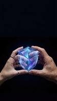 Hands holding glowing blue and purple shield symbol with geometric design on dark background, representing cybersecurity, data protection, and digital safety for tech concepts.