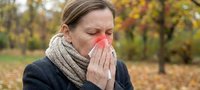 Young Caucasian woman with seasonal cold blowing nose into tissue outdoors in autumn park. Healthcare concept for flu season and respiratory illness prevention.