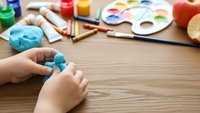 Child hands working with blue modeling clay at wooden table with art supplies, paintbrushes and watercolor palette for creative learning activities.