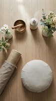 Overhead flat lay of wellness and spa essentials arranged on light wood surface with white flowers, mortar and pestle, diffuser, and round cushion for meditation or relaxation.