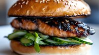 Grilled salmon burger with sesame seed bun, fresh cucumber slices and herbs, glazed with teriyaki sauce, close up view on blurred background.