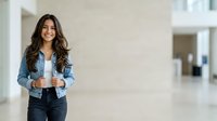 Young woman with long dark hair and warm smile wearing casual denim shirt and dark jeans, standing in modern bright office or corporate environment with neutral beige walls and blurred background space.