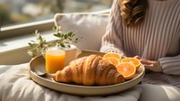 Fresh croissant served with orange juice and citrus slices on ceramic plate in morning sunlight, cozy breakfast setting with woman in sweater near window.