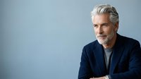 Mature man with gray hair and beard wearing navy blazer over gray shirt, looking at camera with confident expression against neutral blue background.