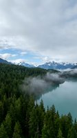 Misty mountain lake surrounded by dense evergreen forest with snow-capped peaks under dramatic cloudy sky creating serene wilderness landscape.