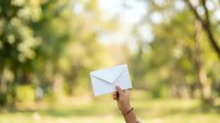 Hand holding white envelope outdoors in sunny park setting with blurred green trees, representing mail delivery, correspondence, communication, and messaging concepts.