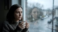 Young woman with dark hair and fair complexion holding warm beverage while gazing through rainy window, contemplative mood, suitable for lifestyle, wellness, seasonal and emotional storytelling content.