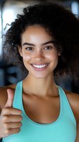 Young mixed race woman with curly hair giving thumbs up gesture wearing mint green tank top smiling at camera in fitness gym environment for wellness motivation.