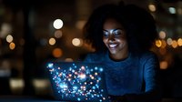 Young African American woman smiling while interacting with glowing holographic display in dark room with bokeh lights, creating magical technology atmosphere.