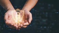 Hands holding glowing digital security shield with lock icon over dark circuit board background representing cybersecurity protection and data safety.