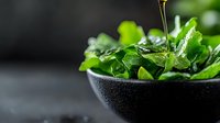 Fresh green watercress leaves in black bowl with olive oil drizzle on dark background, macro food photography with selective focus and water drops.