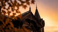 Victorian Gothic architecture house with ornate wooden details and spires silhouetted against warm sunset sky with tree branches framing the scene.