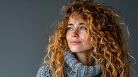 Young woman with curly blonde hair wearing cozy gray knit sweater looking up thoughtfully against neutral background for lifestyle content.