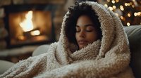Young African American woman wrapped in cozy sherpa blanket relaxing by fireplace with Christmas lights in background, eyes closed in peaceful winter moment.