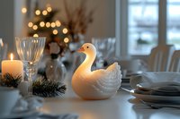 Elegant white ceramic swan centerpiece on festive holiday dining table with candles, pine branches, crystal glasses and warm bokeh lights creating cozy atmosphere.
