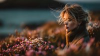 Young blonde woman enjoying peaceful moment among purple heather flowers during golden hour sunset with warm natural lighting and serene outdoor atmosphere.