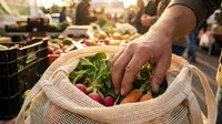 Man holding mesh bag filled with fresh vegetables at farmers market, displaying locally grown produce including radishes and carrots for sale.