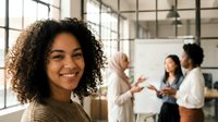 Smiling young African American businesswoman in modern office with diverse team collaborating on presentation in background workspace.