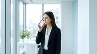 Professional businesswoman drinking coffee in modern office with large windows. Young female executive in black blazer enjoying morning beverage break at workplace.