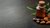 Amber glass bottle with cork stopper containing almond oil surrounded by fresh almonds and green leaves on dark slate surface for natural skincare and aromatherapy.