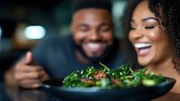 Young African American couple sharing joyful moment over fresh healthy salad plate in romantic restaurant setting, focus on green leafy dish in foreground.