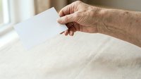 Senior adult male hand holding blank white card over neutral fabric surface with natural window lighting.
