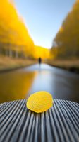 Yellow autumn leaf on wooden bench with blurred figure walking along canal path amid golden fall foliage and blue sky reflection in water.